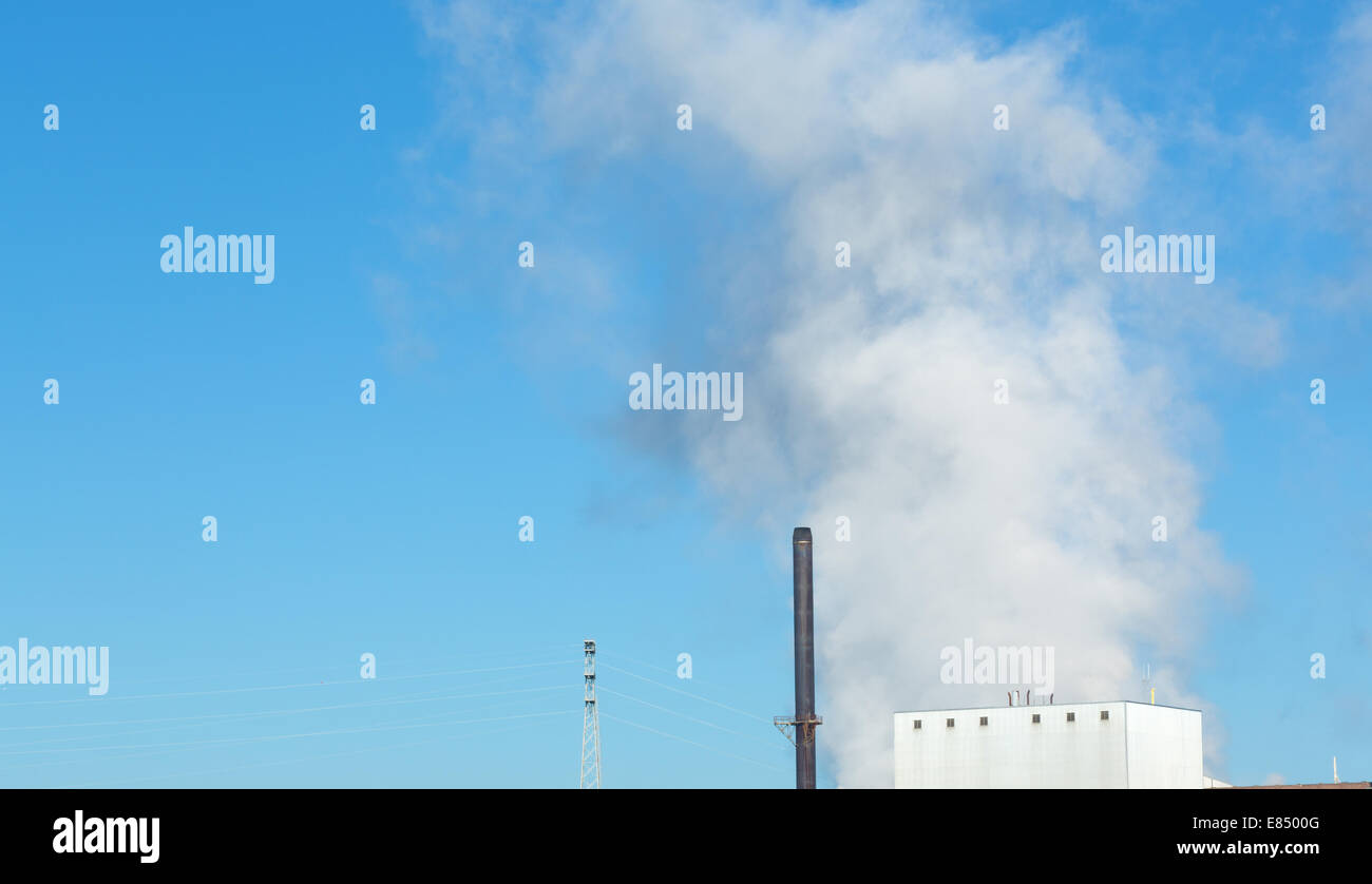 A large emission of steam rising from a factory against a bright blue sky with building and smokestacks in the foreground. Stock Photo