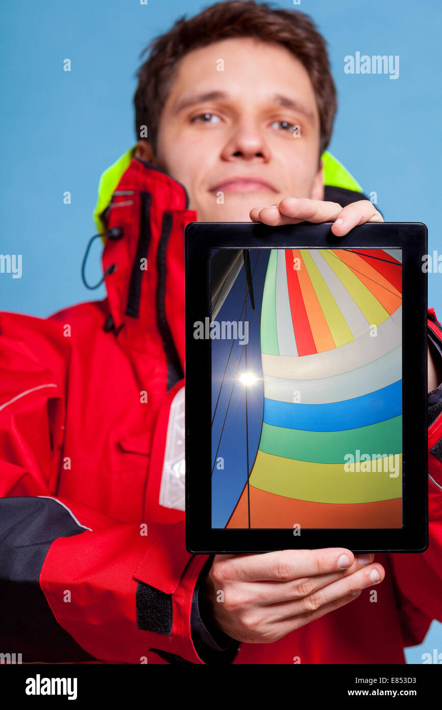 Man in wind jacket holding ipad with photo of colorful sail of boat. Sailor showing screen tablet touchpad dreaming about yacht Stock Photo