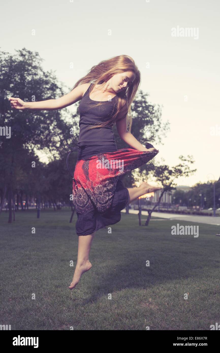 Long Haired Blonde  jumps on a green grass in evening time in city park . Girl jumping like flying bird. Stock Photo