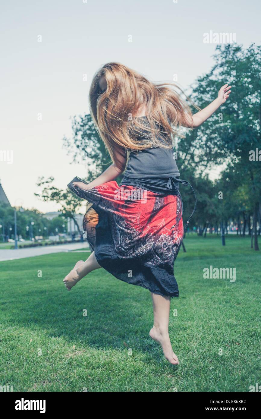 The long haired 20s women jumps on a green grass in evening time in city park . Girl jumping like flying bird. Stock Photo