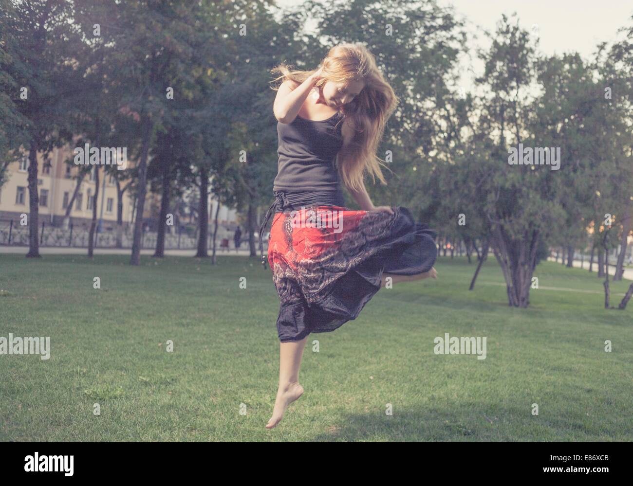 The young female jumps on a green grass in evening time in city park . Girl jumping like flying bird. Stock Photo