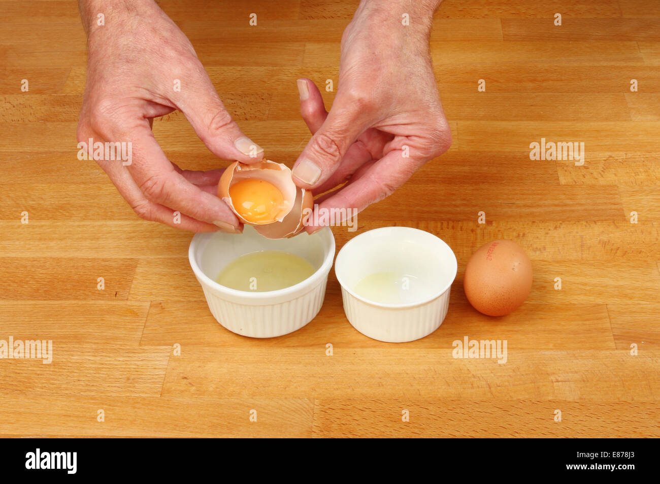 Hands separating egg yolks into ramekins on a wooden worktop Stock Photo
