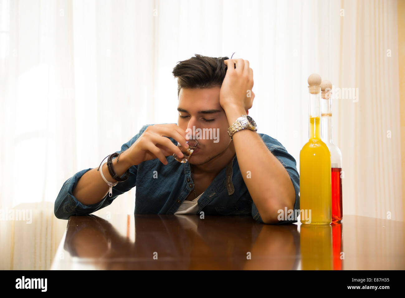 Sleepy, drunk young man sitting drinking alone at a table with two bottles of liquor alongside him sipping from shot glass to Stock Photo
