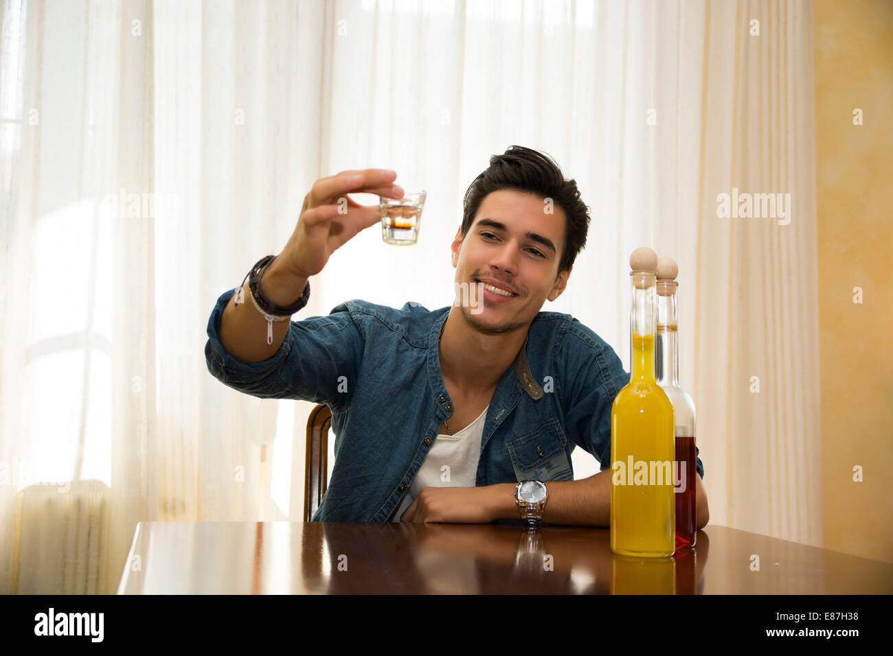 Young man sitting drinking alone at a table, making a toast, with two bottles of liquor alongside him sipping from shot glass Stock Photo