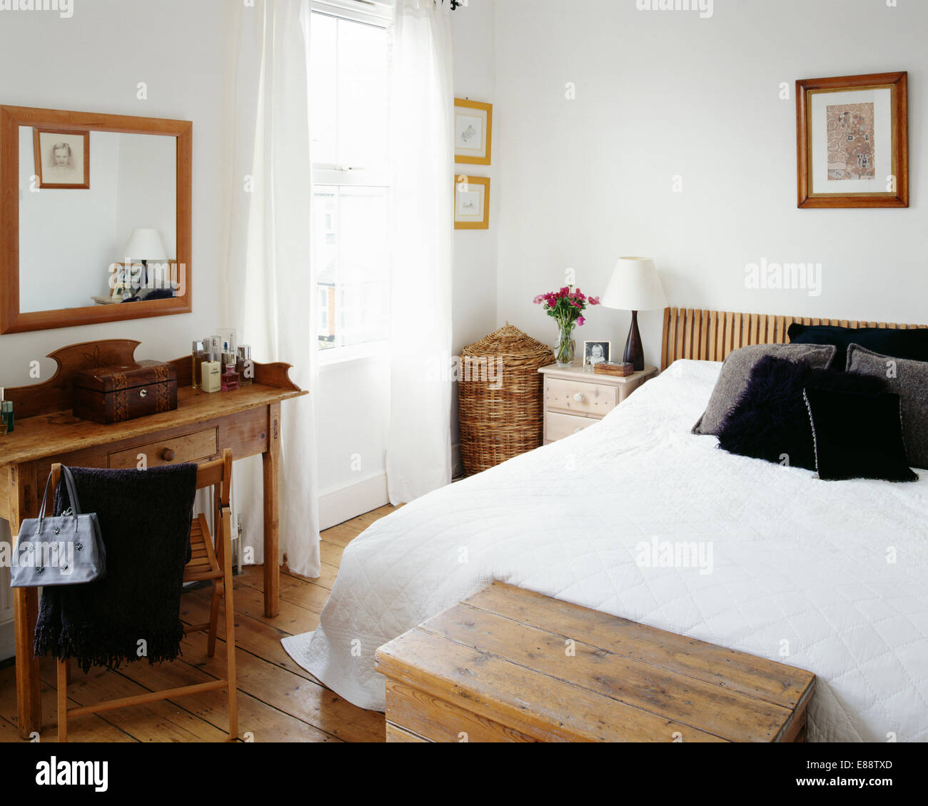White quilt and black cushions on bed in economy-style bedroom with wooden mirror above simple pine table Stock Photo