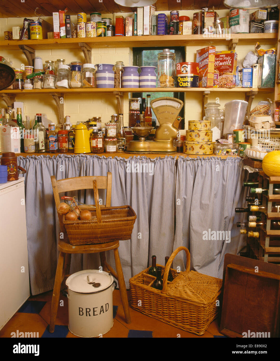 Storage jars on wooden shelves above scales on worktop with curtain in larder with baskets and