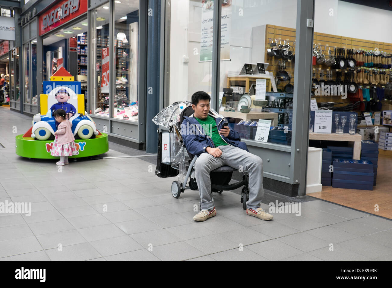 A bored Asian father sitting in his daughters buggy, playing with his smart phone & not watching his child whilst shopping Stock Photo