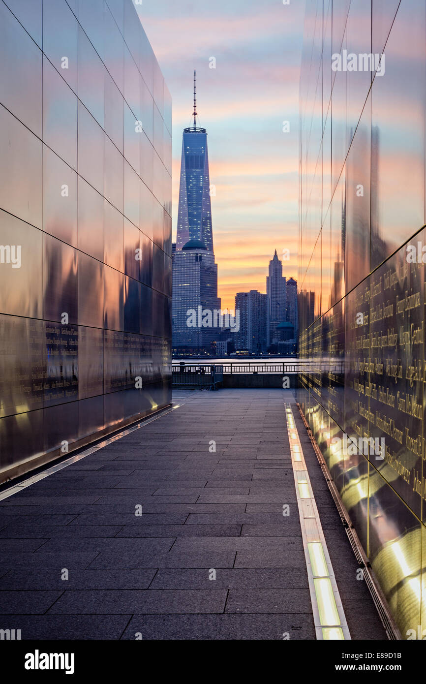 The colors of sunrise reflect on the walls of the Empty Sky Memorial at Liberty State Park with the Freedom Tower at One World Trade Center in the Financial District in New York City. Stock Photo