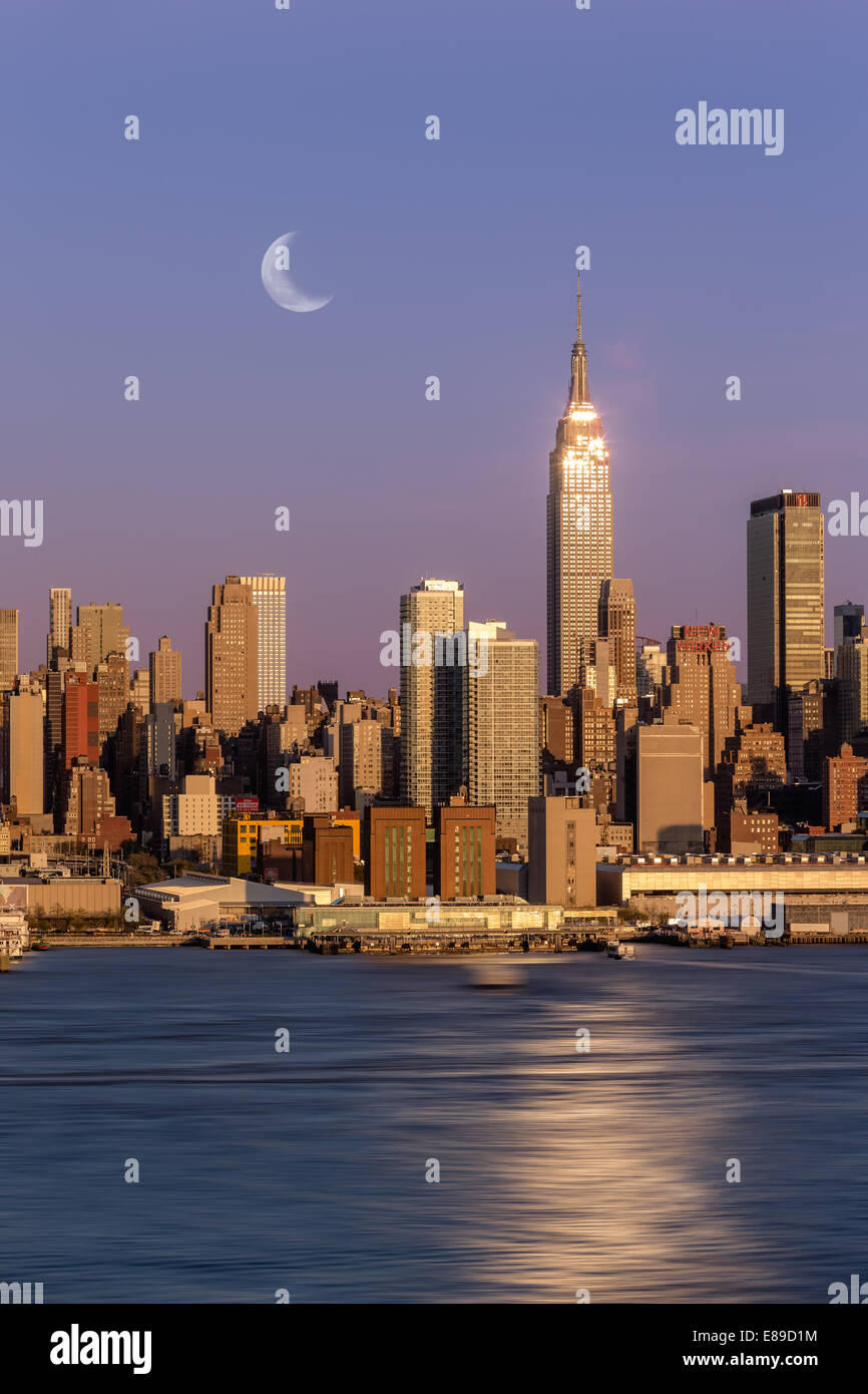 New York City skyline with the Empire State Building brightly lit from the setting sun and a half moon as well. Stock Photo