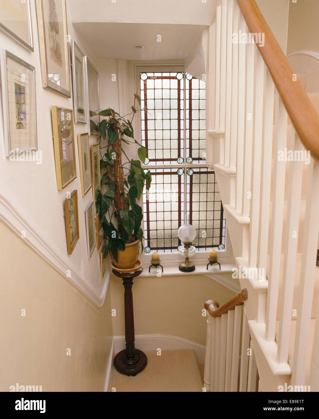 Trailing houseplant in pot on wooden pedestal beside stained glass window above stairs with white banisters Stock Photo