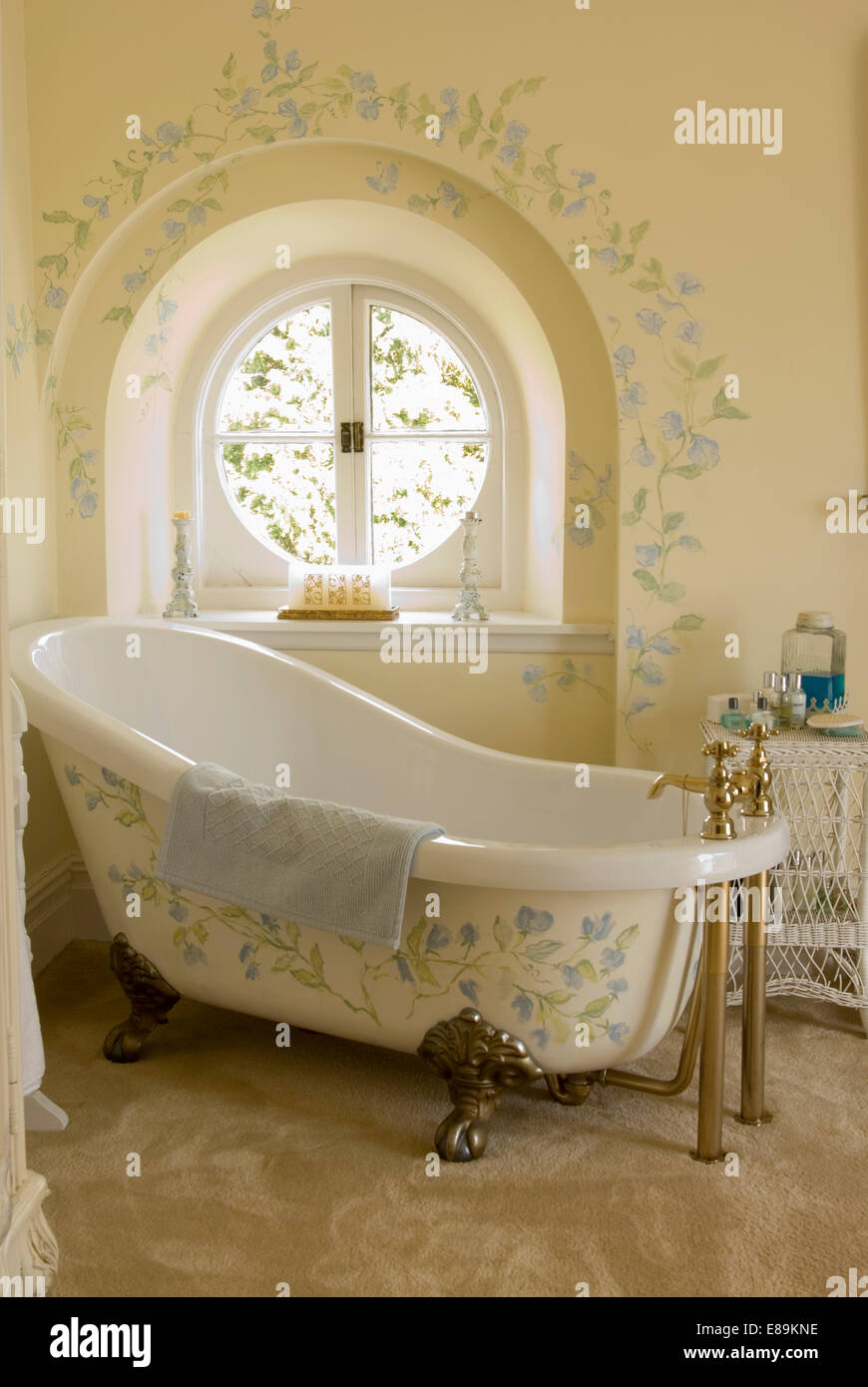 Cream bathroom with painted floral decoration on rolltop bath in front of circular window with matching decoration on wall Stock Photo