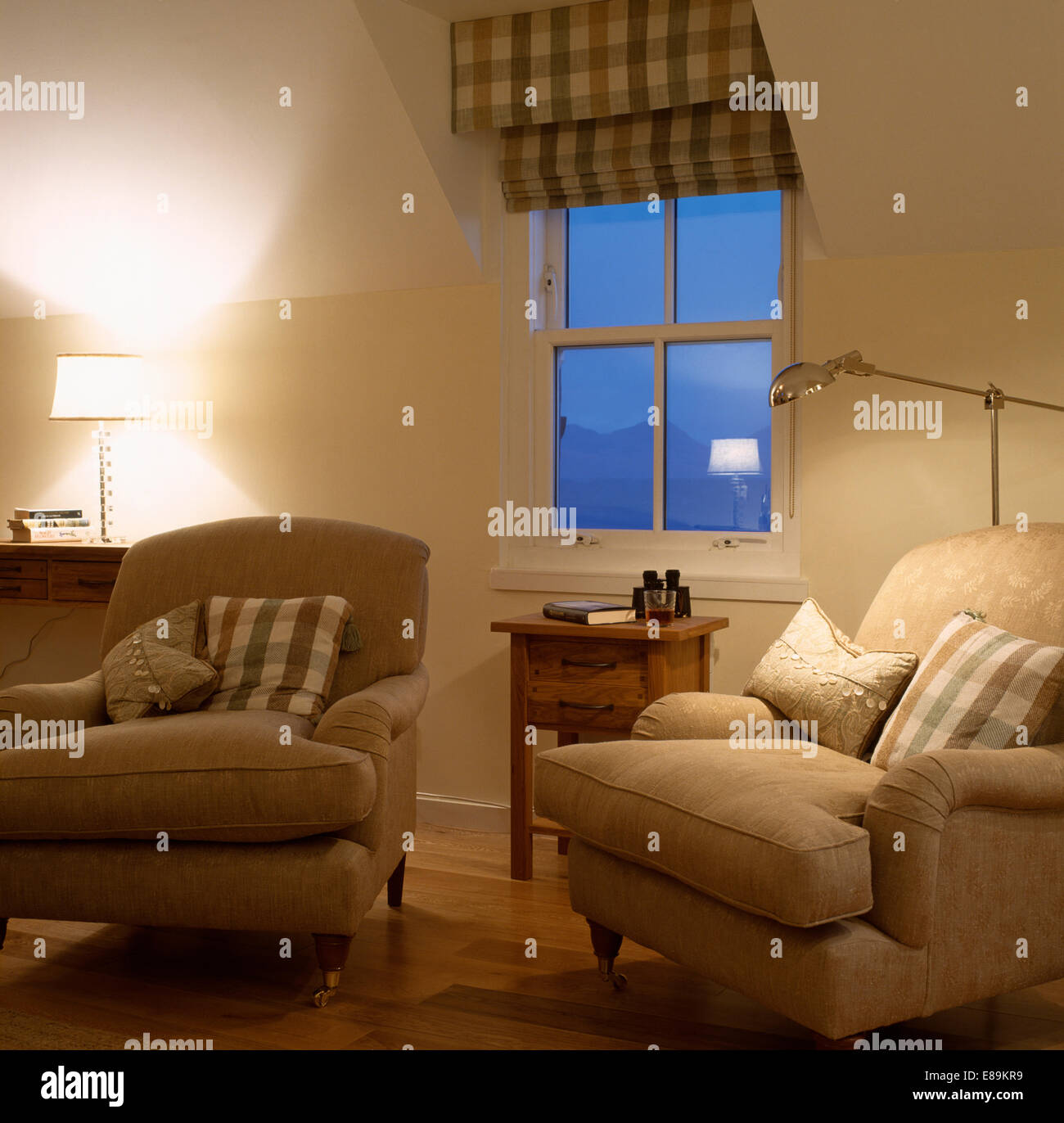 Checked blind on window in cream coastal living room with lighted lamps and cream armchairs Stock Photo