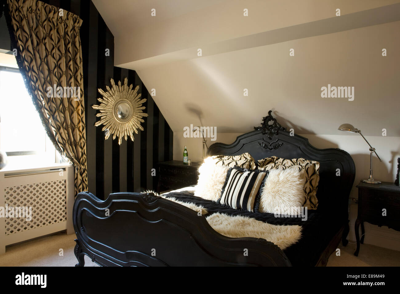 Period style black bed with faux fur cushions in loft conversion bedroom with black+gold patterned curtain Stock Photo