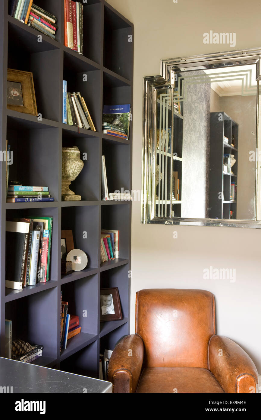Art deco style mirror above leather armchair in corner of modern study with painted shelving Stock Photo