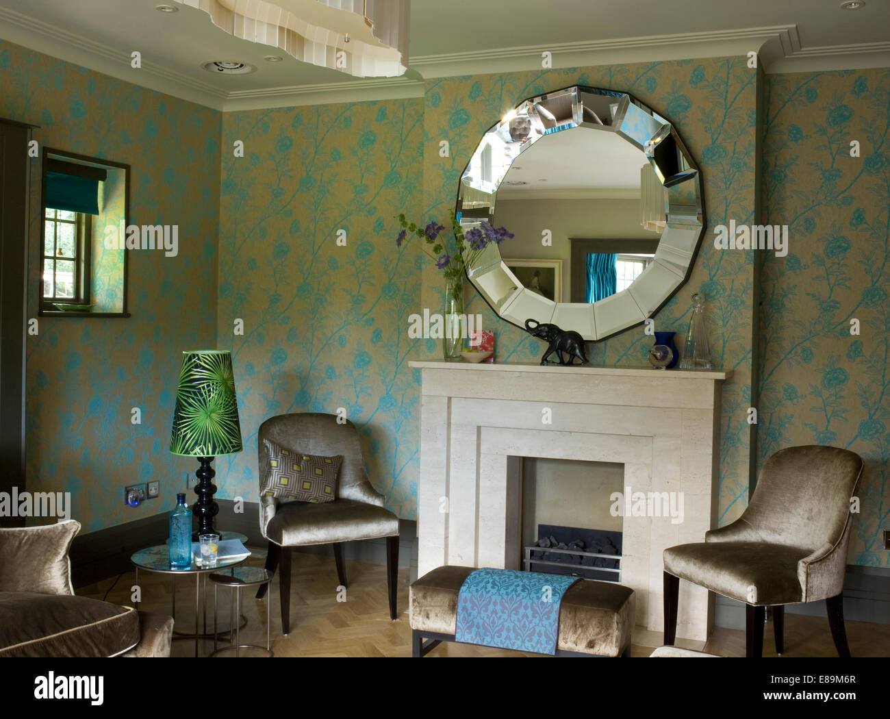 Round mirror above fireplace in traditional sitting room with grey velvet upholstered chairs and stool Stock Photo