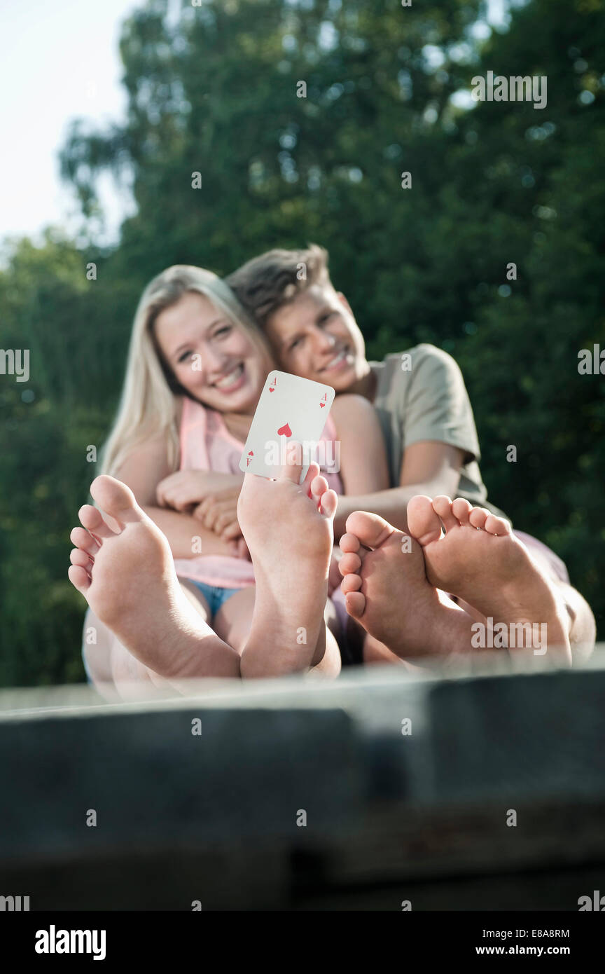 Teenage couple sitting on a jetty at lake, girl holding an ace between her toes Stock Photo