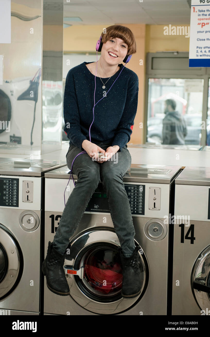 Young woman listening music on top of washing machine, smiling Stock