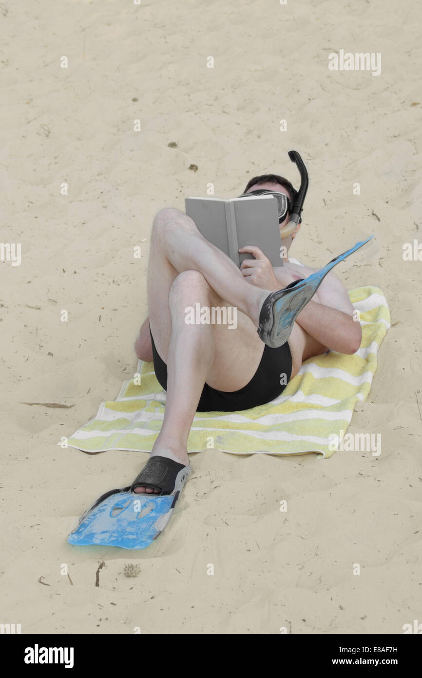 Man in flippers and diving mask with snorkel lays on sand and reads book Stock Photo