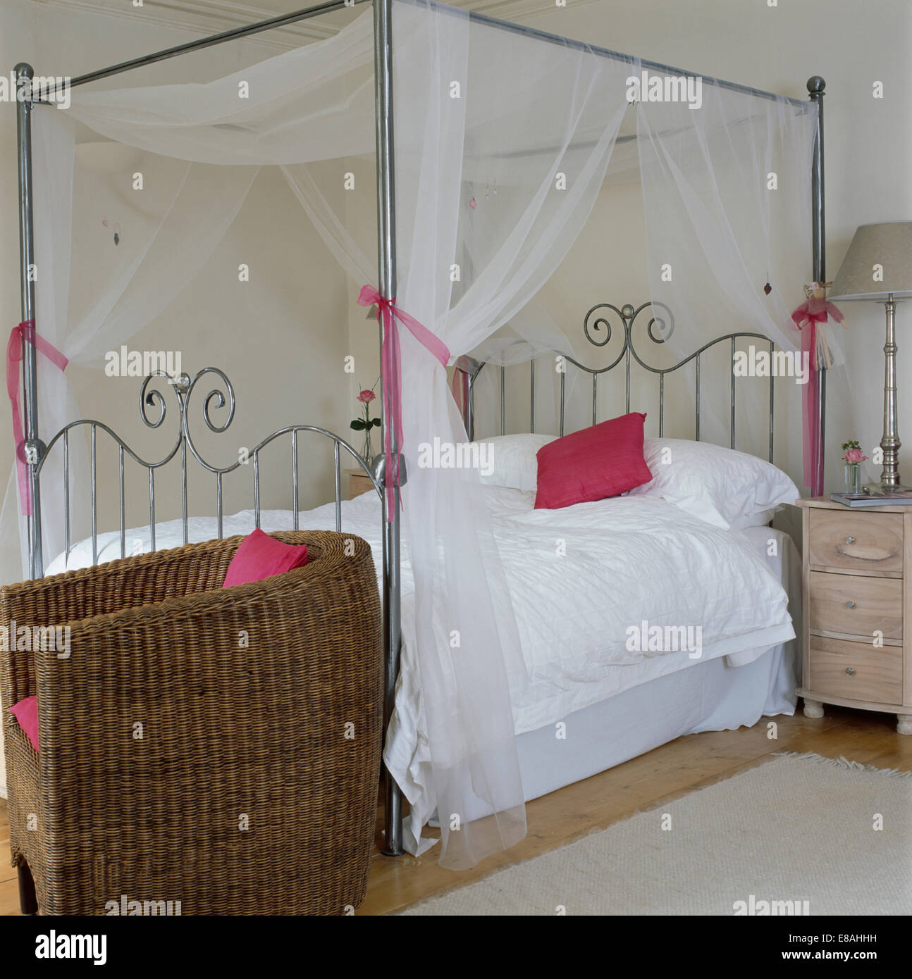 Wicker tub chair beside chrome four-poster bed with white voile drapes and white duvet in white country bedroom Stock Photo