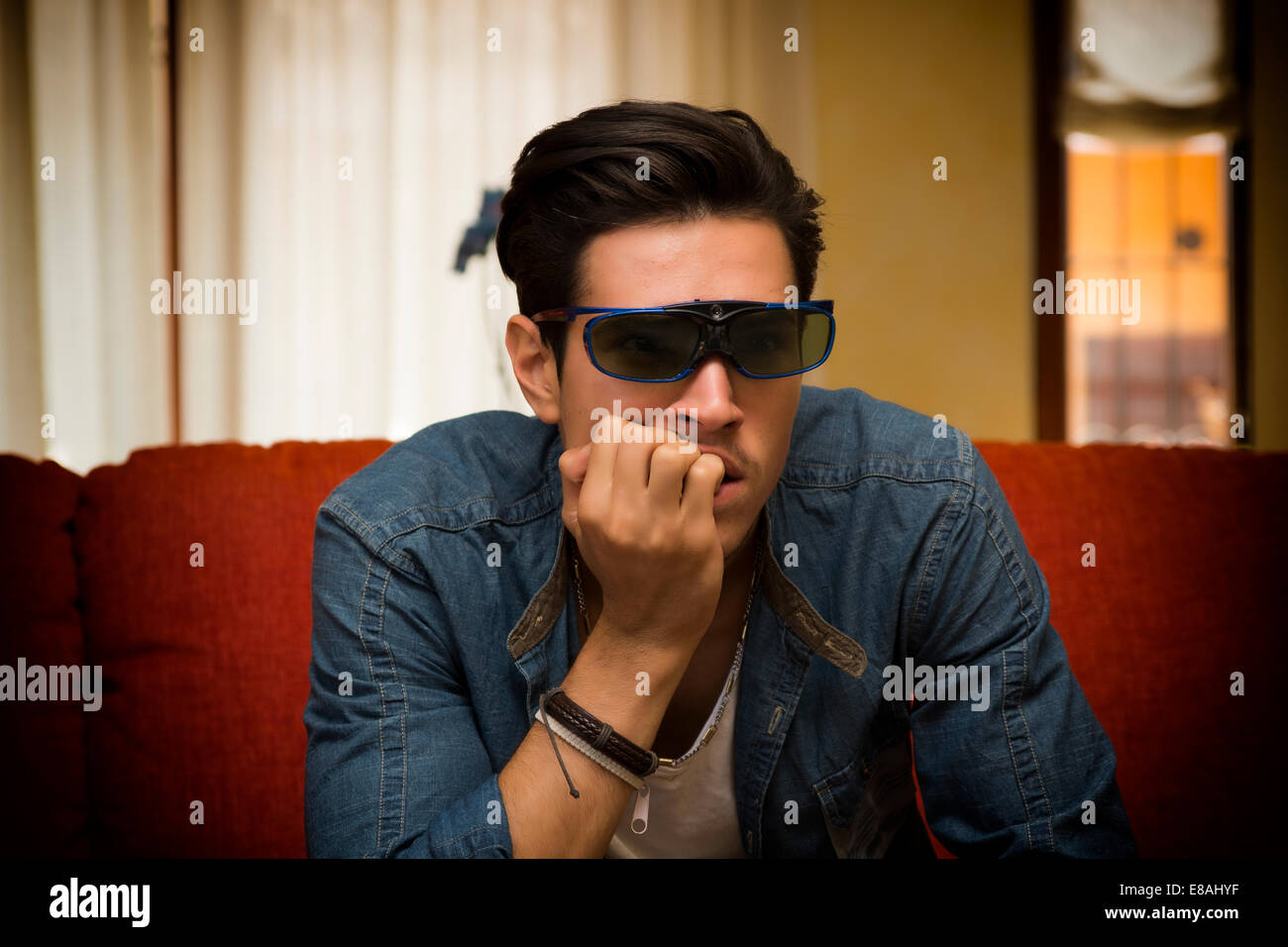 Young man wearing stereoscopic 3d glasses sitting watching a video or television program on a sofa at home sitting forwards with Stock Photo