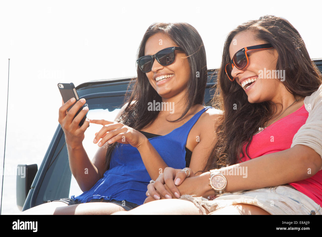 Two young women sitting on jeep hood looking at smartphone Stock Photo