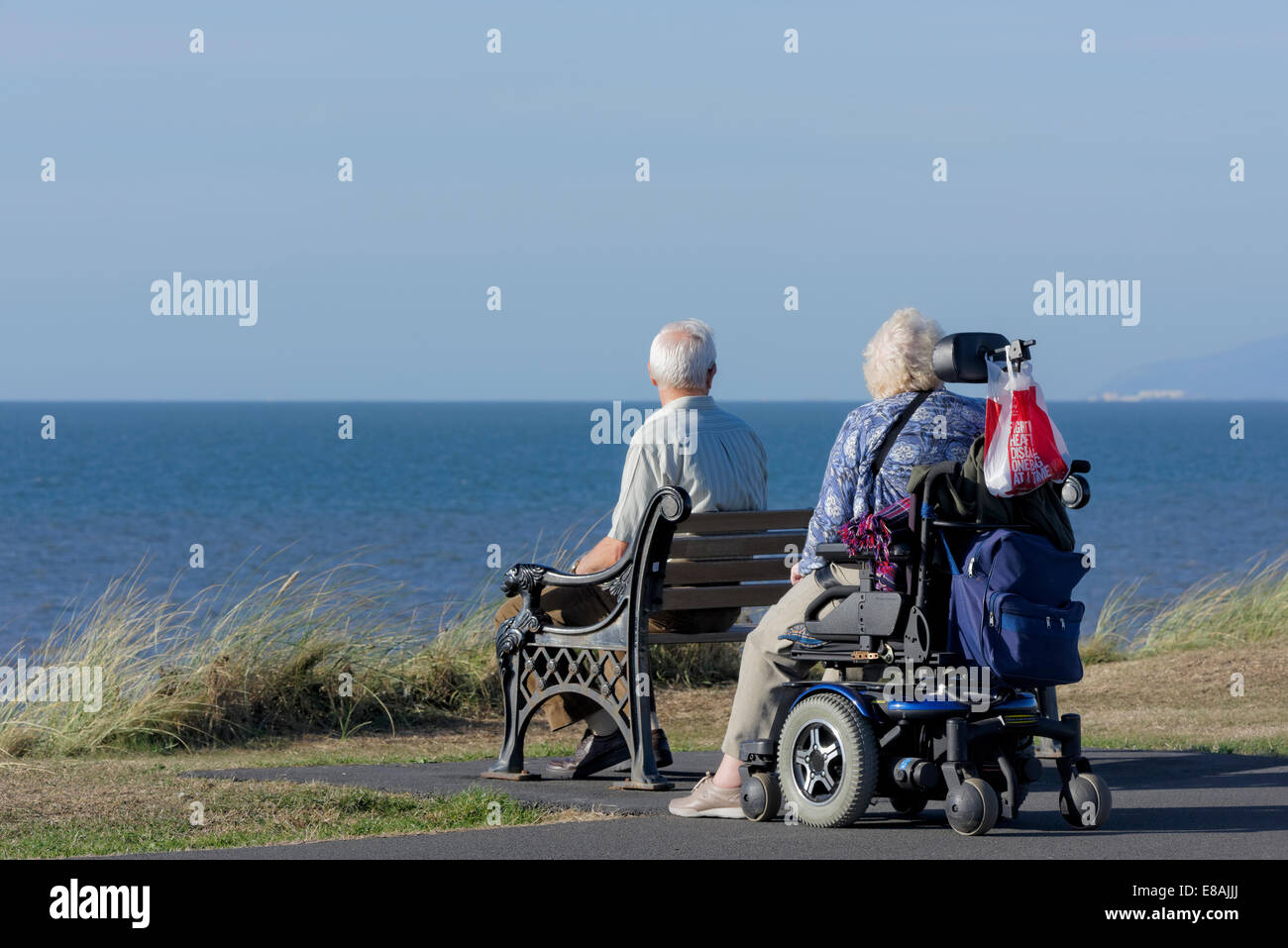 Man sitting on bench with wife in wheelchair on a coastal path in Blackpool, Lancashire, UK Stock Photo