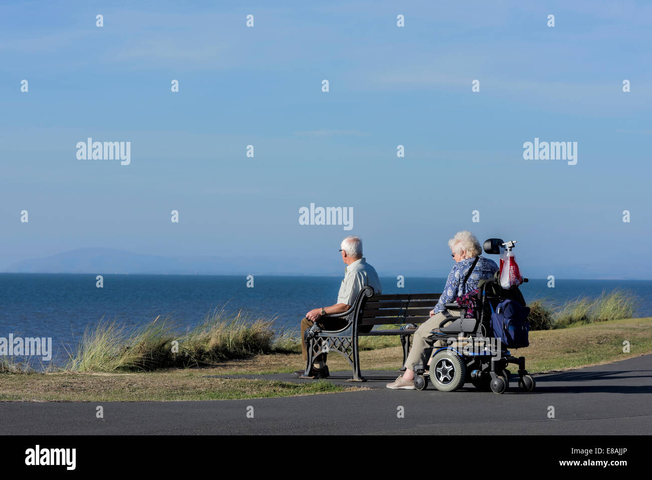 Man sitting on bench with wife in wheelchair on a coastal path in Blackpool, Lancashire, UK Stock Photo