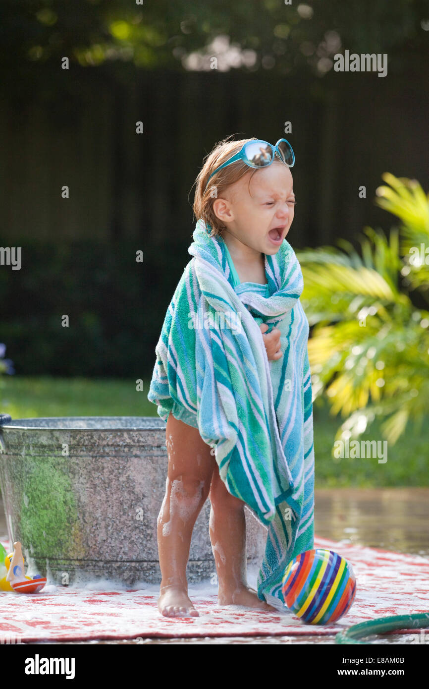 Female toddler wrapped in towel crying beside bubble bath in garden
