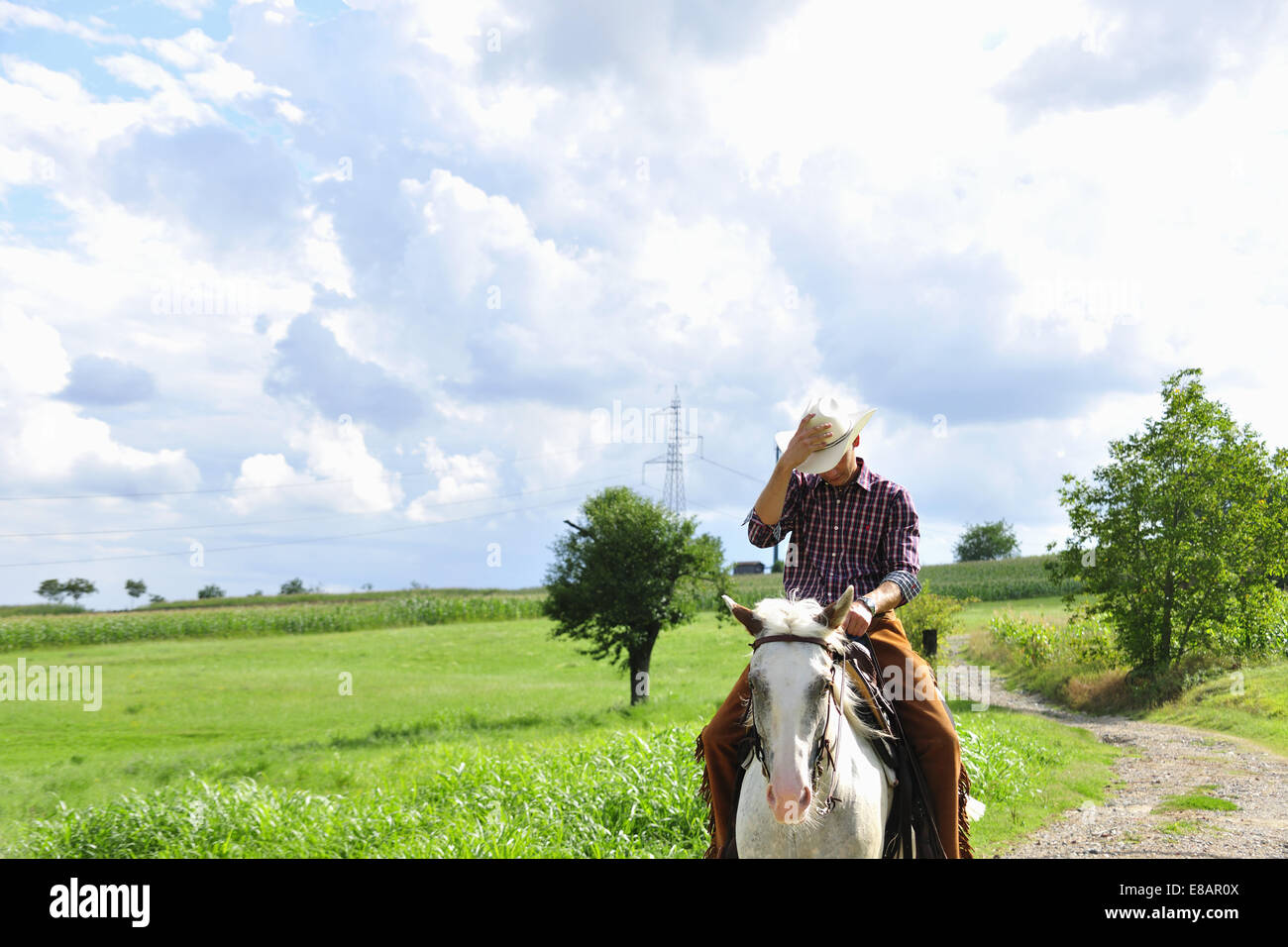 Young man adjusting cowboy hat riding horse on rural road Stock Photo