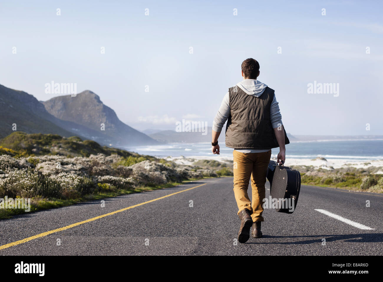 Rear view of young man with guitar case walking on coastal road, Cape Town, Western Cape, South Africa Stock Photo