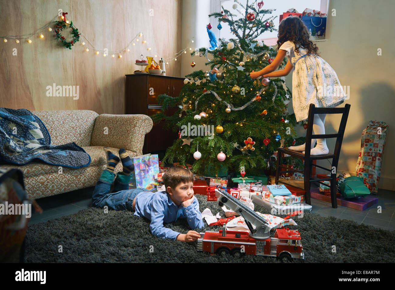 Girl arranging christmas tree whilst brother plays with xmas gifts on sitting room floor Stock Photo