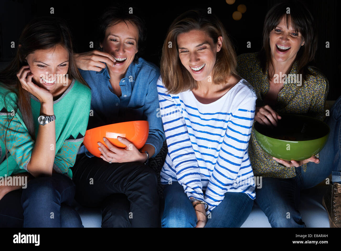 Four women laughing and watching TV with snack bowls Stock Photo
