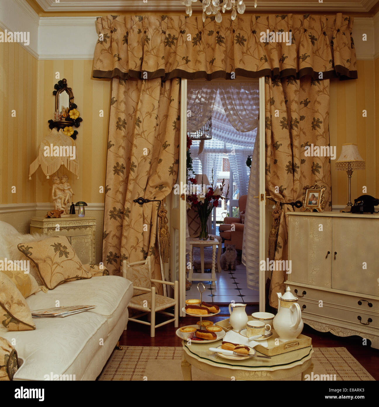 Tea set on tray in small living room with concealed television and patterned curtains at open French windows to conservatory Stock Photo