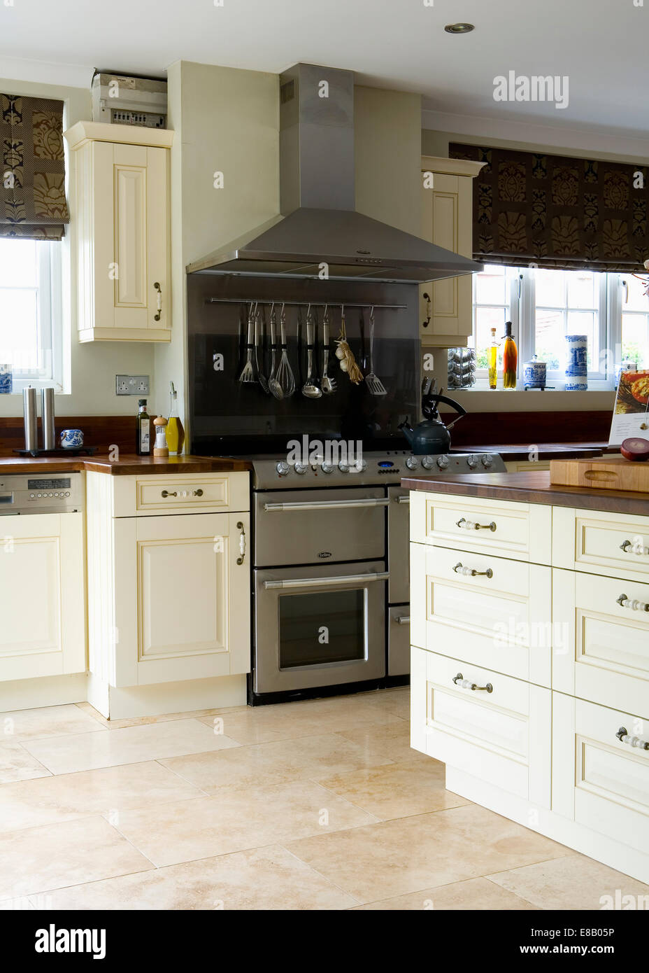 Cream travertine floor tiles in modern kitchen with large stainless steel range oven and cream fitted units Stock Photo