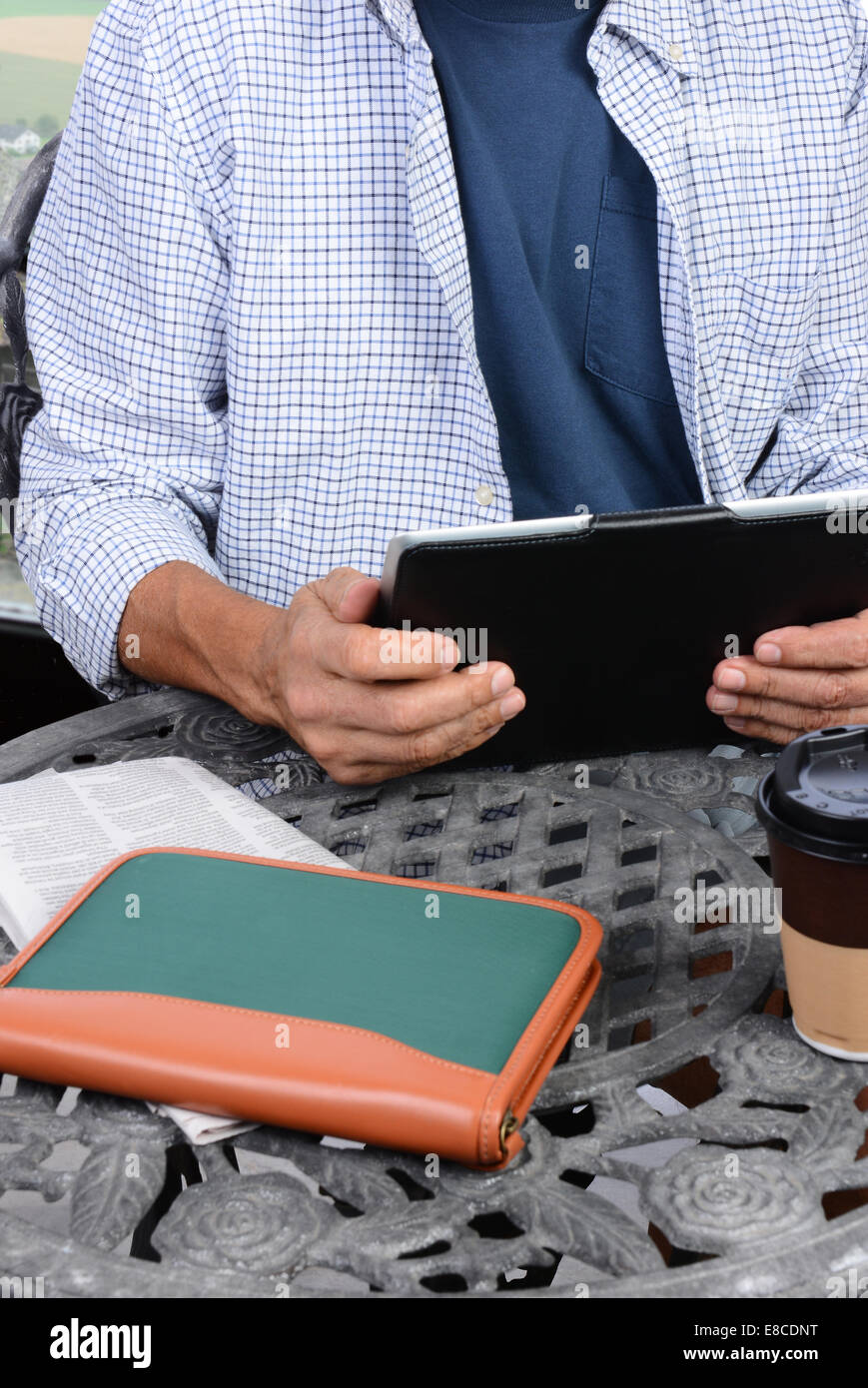 Closeup of a man using his tablet computer an the table of an outdoor internet cafe. A newspaper, coffee cup and notebook are on Stock Photo