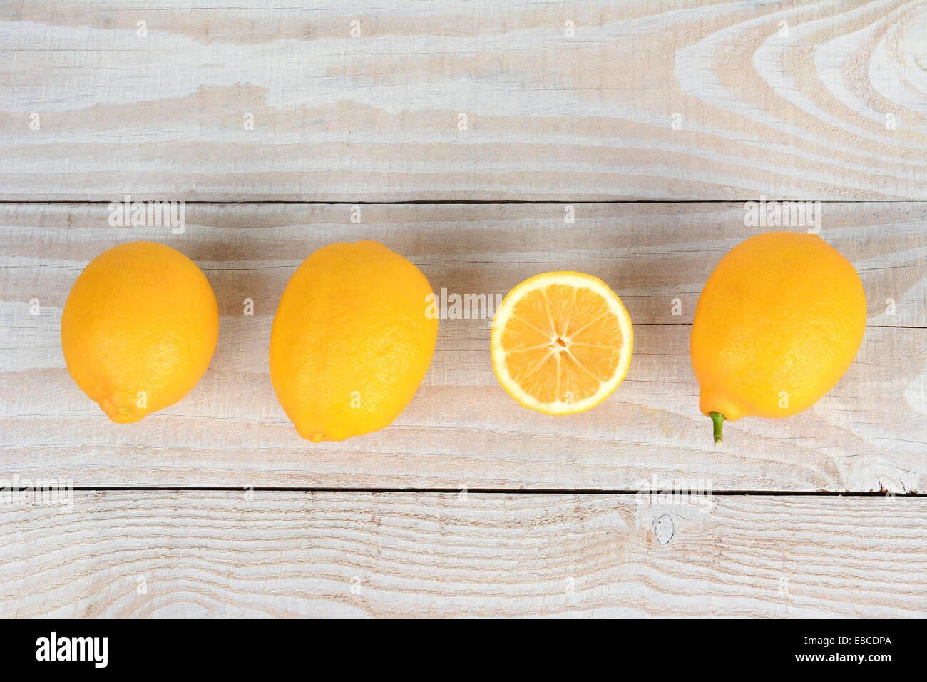 A row of lemons on a white wood kitchen table. One of the lemons is cut in half exposing is flesh. Stock Photo