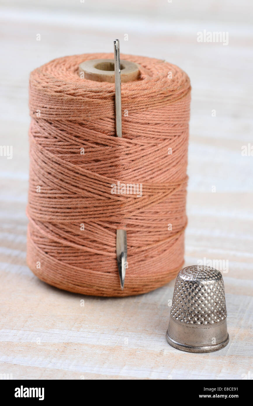 Closeup of an old thimble, large needle and spool of thread on a white wood background. Focus is on the thimble. Stock Photo