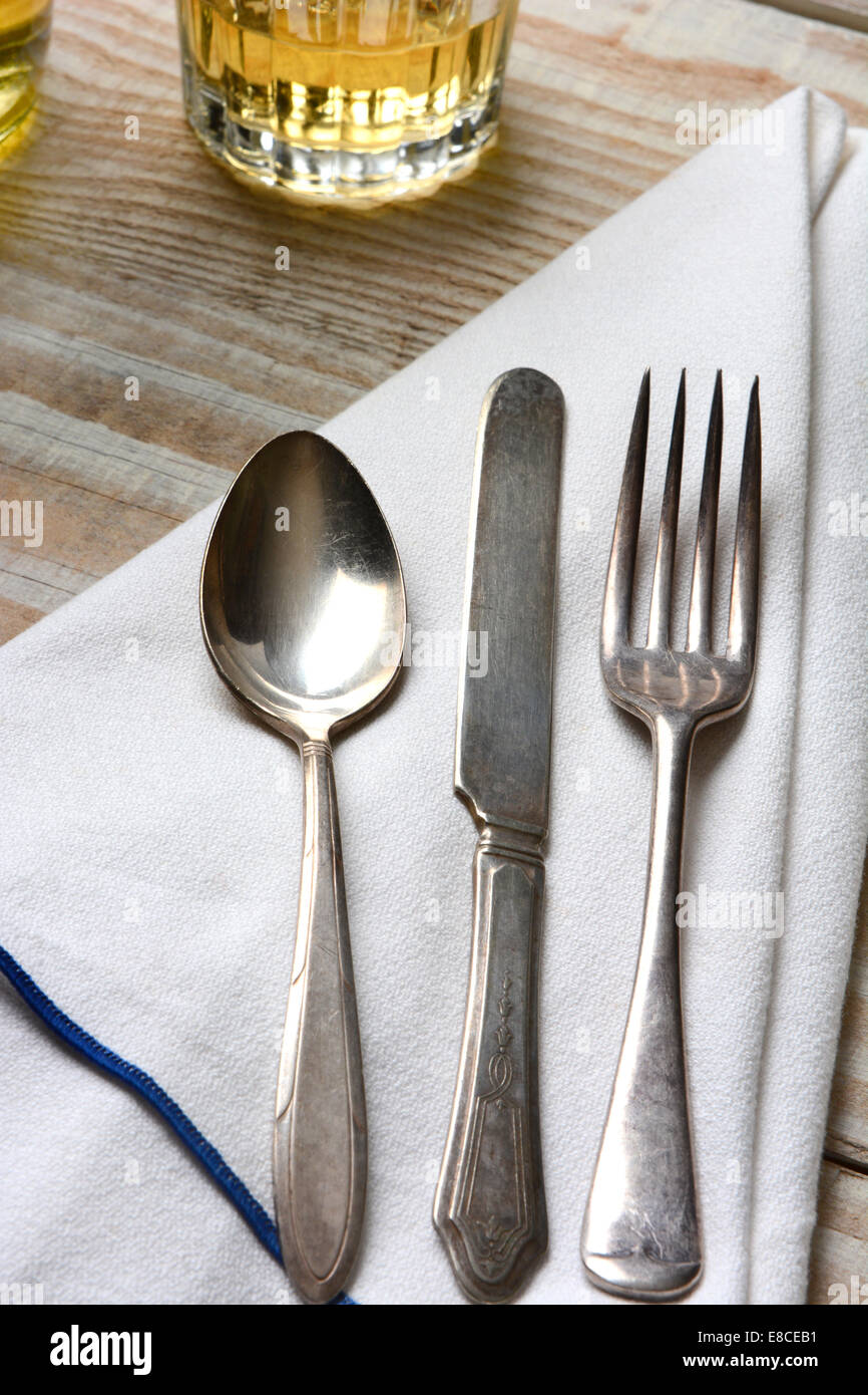 Closeup of a table setting. Old mismatched silverware, napkin and drinks on a white wood table. Vertical format. Stock Photo