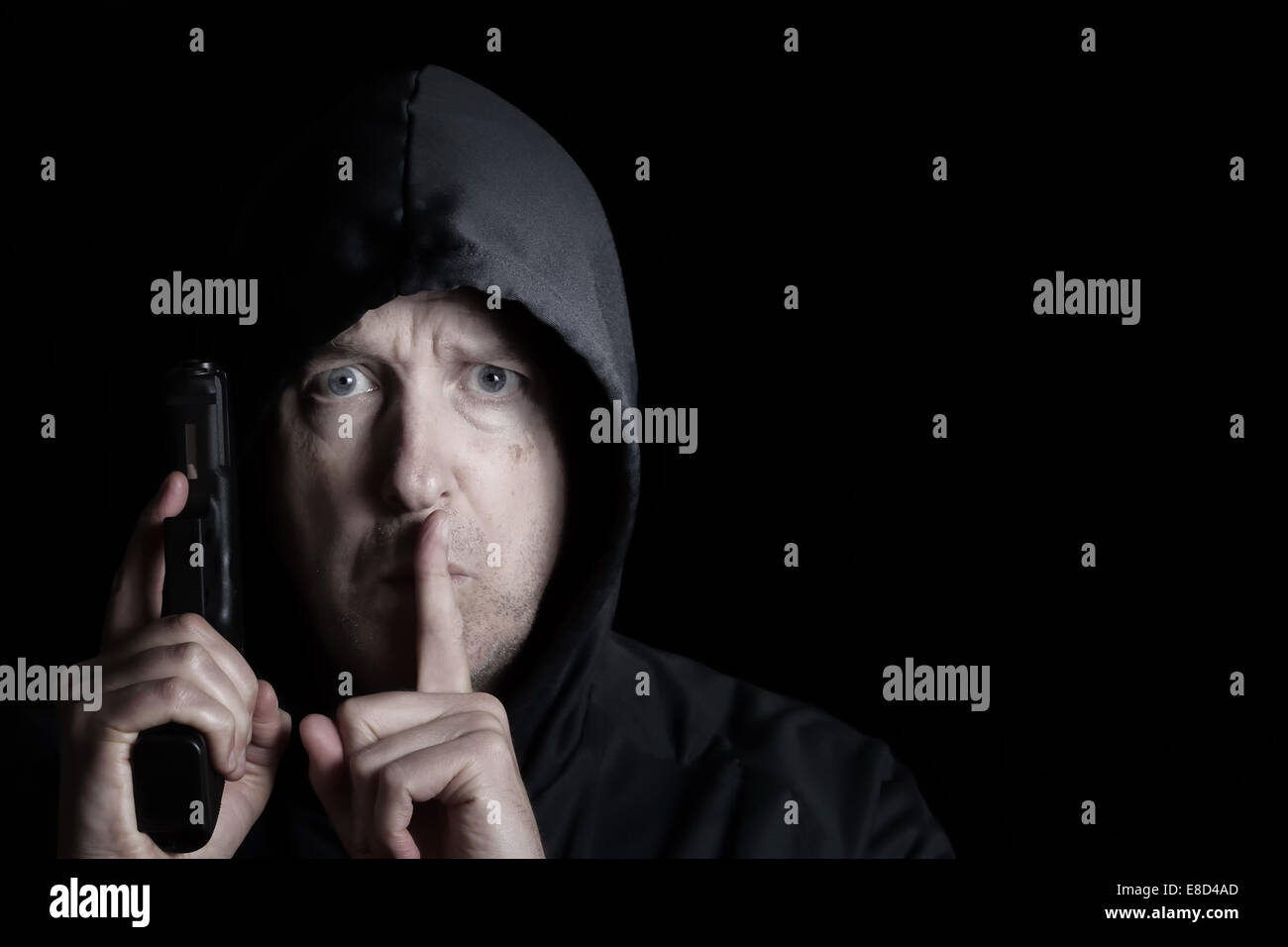 Closeup front view of mature man, looking forward and wearing hood, with weapon and index finger signaling to be silent on dark Stock Photo