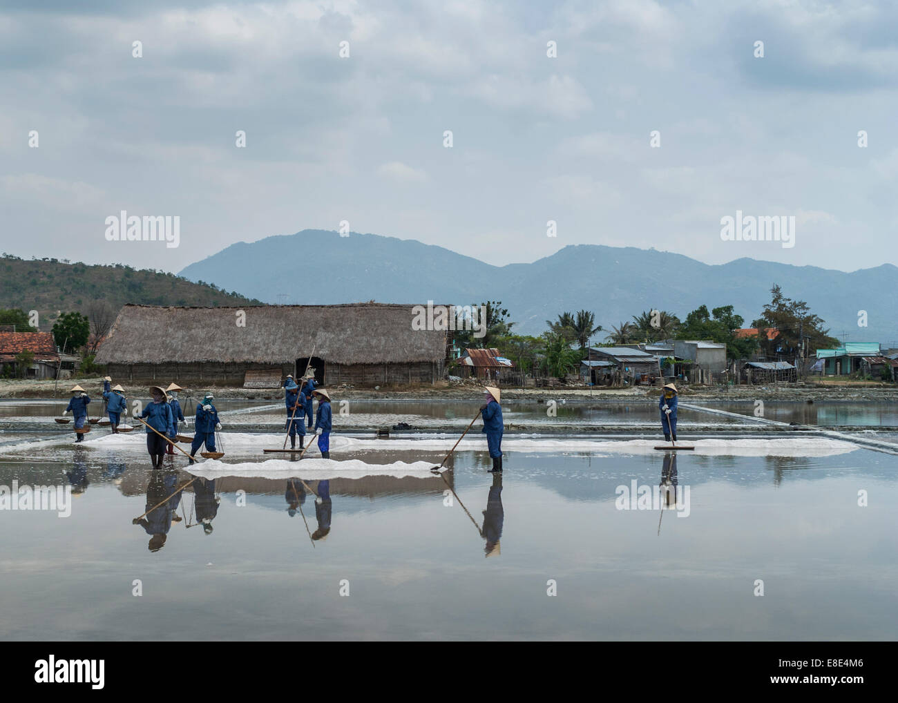 South Vietnam - March 2012: Crew of blue-clad people rake, scoop and carry baskets of freshly harvested salt. Stock Photo
