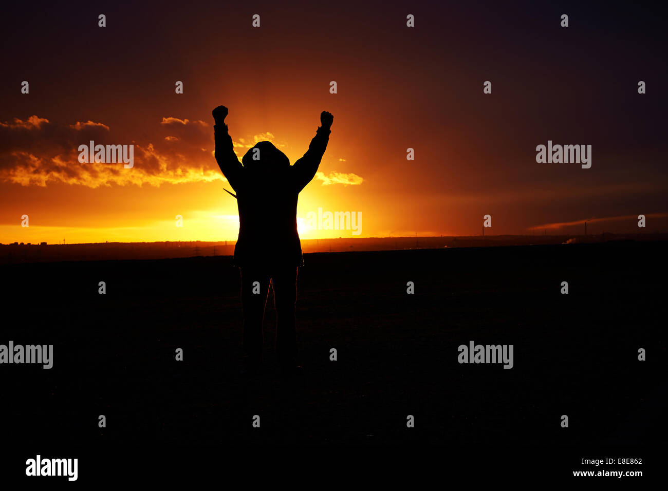 Silhouette of a man holding up his arms in jubilation behind a dark orange colored sunset. Long shot. Stock Photo