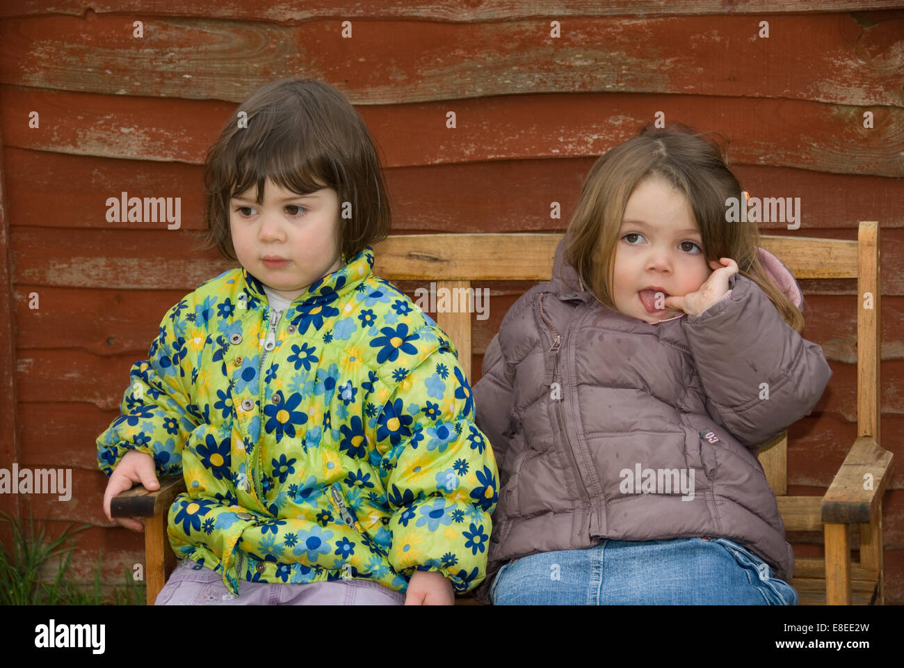 2 toddler girls wearing anoraks aged 3 4 5 on a bench with a shed background Stock Photo