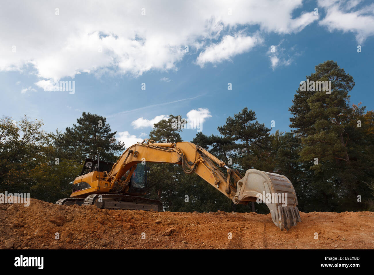 side view of excavator on construction site outdoors, with forest and blue clouded sky in background Stock Photo