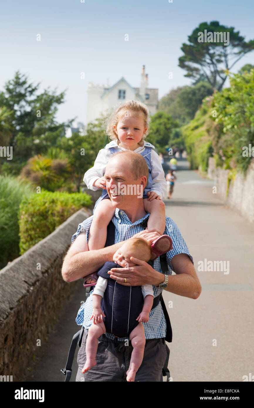Mature dad / father with two / 2 children; one child on shoulders and baby In sling. Cornwall UK. Stock Photo