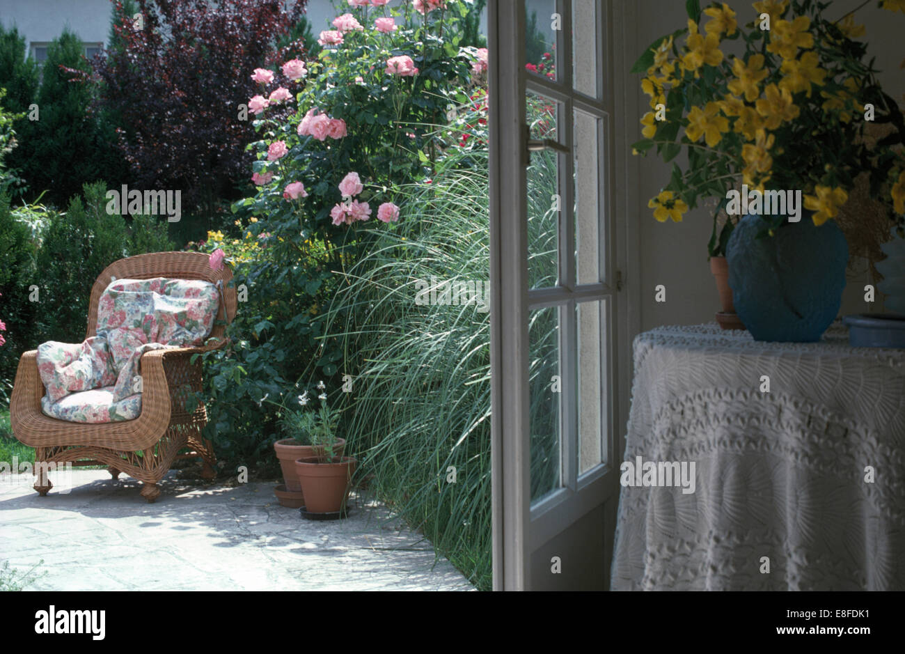 Yellow flowers in vase on table beside open doors to terrace with a wicker armchair beside pink climbing roses Stock Photo