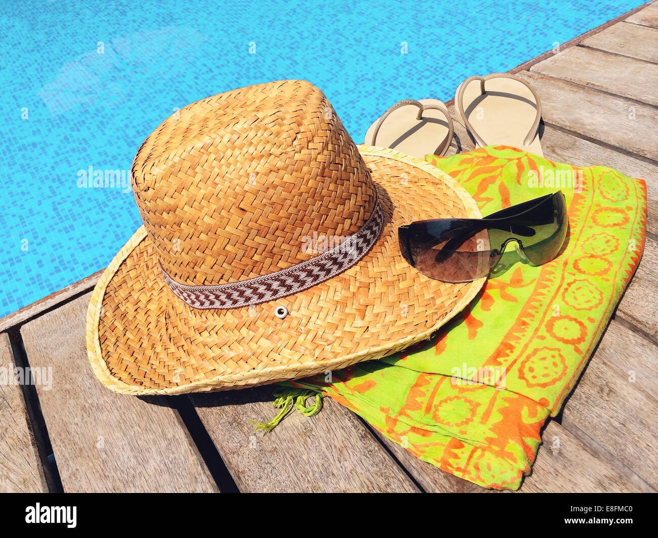 Straw hat, sunglasses, flip flops and sarong on wooden deck by swimming pool, Thailand Stock Photo