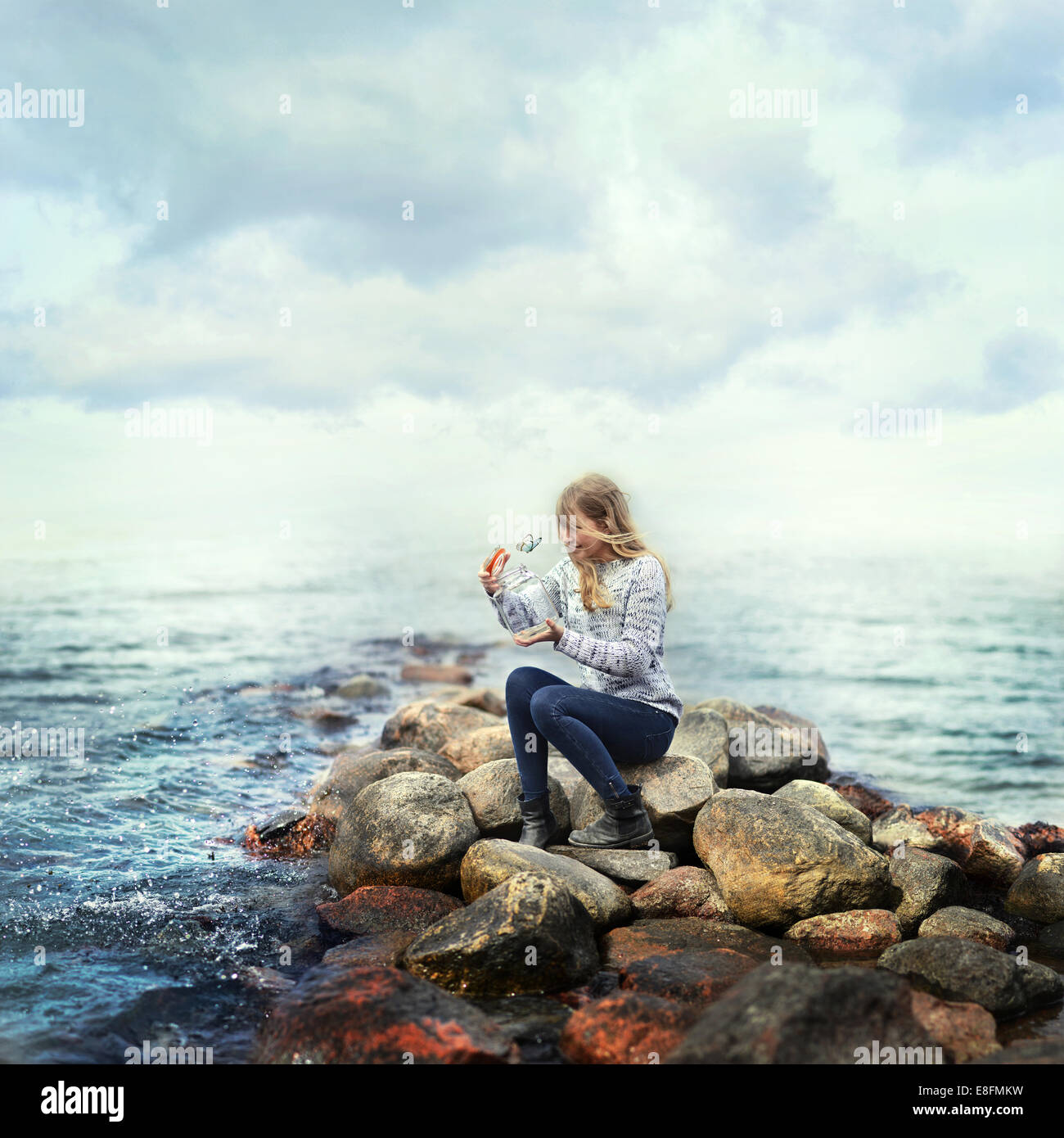 Woman sitting on rocks by ocean releasing a butterfly from a glass jar ...