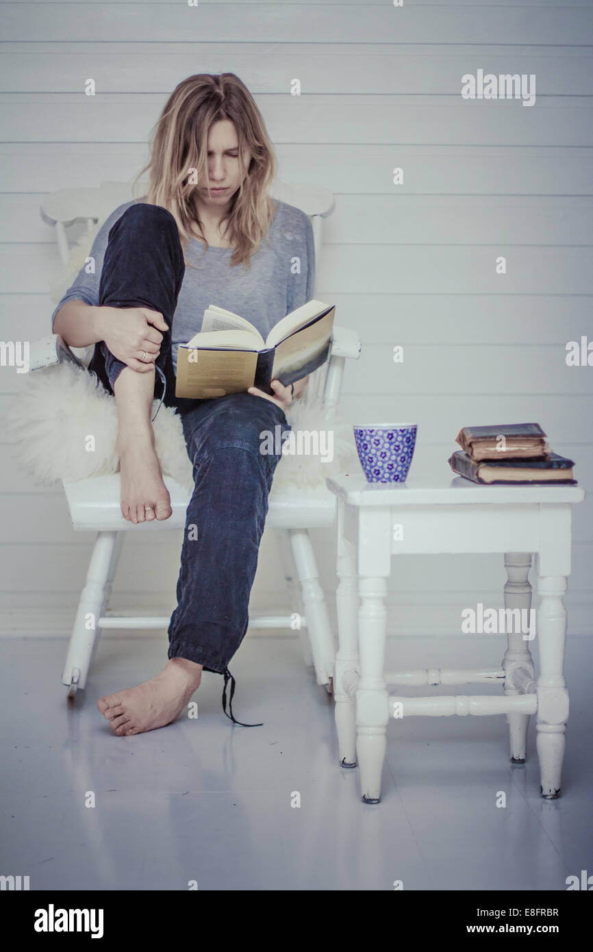 Portrait of a woman sitting on chair reading a book with cup of tea Stock Photo