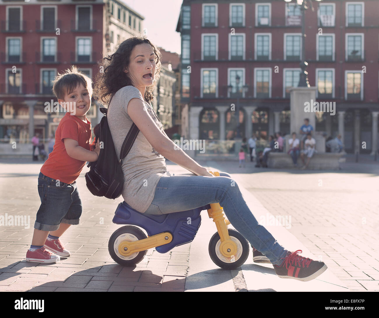 Boy pushing his mother on a toy tricycle Stock Photo