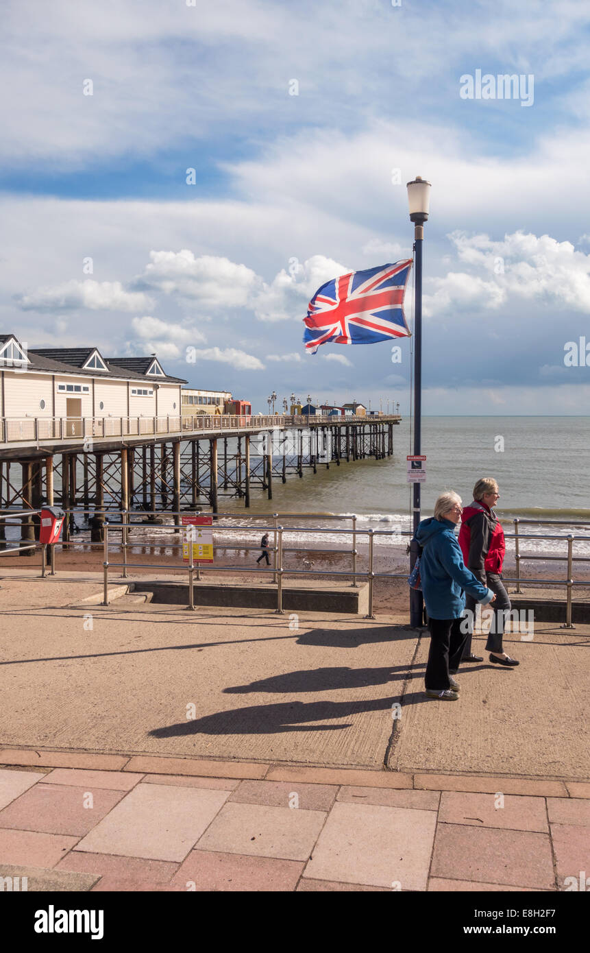 The pier stretching out to sea at Teignmouth in Devon with a union jack fluttering in the foreground. Two women walking together Stock Photo